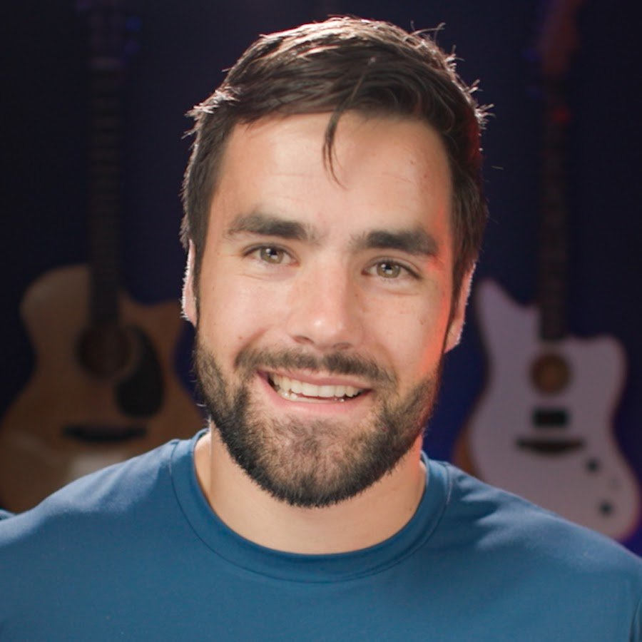 A smiling man with dark brown hair and a beard, wearing a blue T-shirt, in front of a background with multiple guitars hanging on the dark wall.